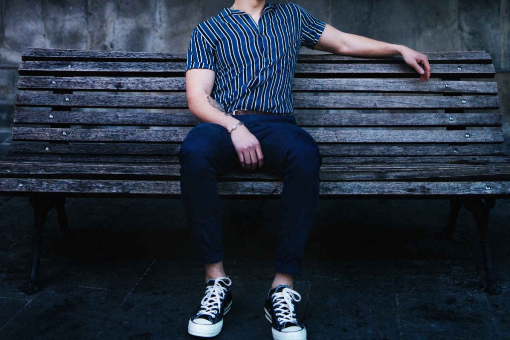 Young man sitting on a park bench, wearing a blue striped short sleeve shirt, blue pants and a pair of black Converse shoes without socks