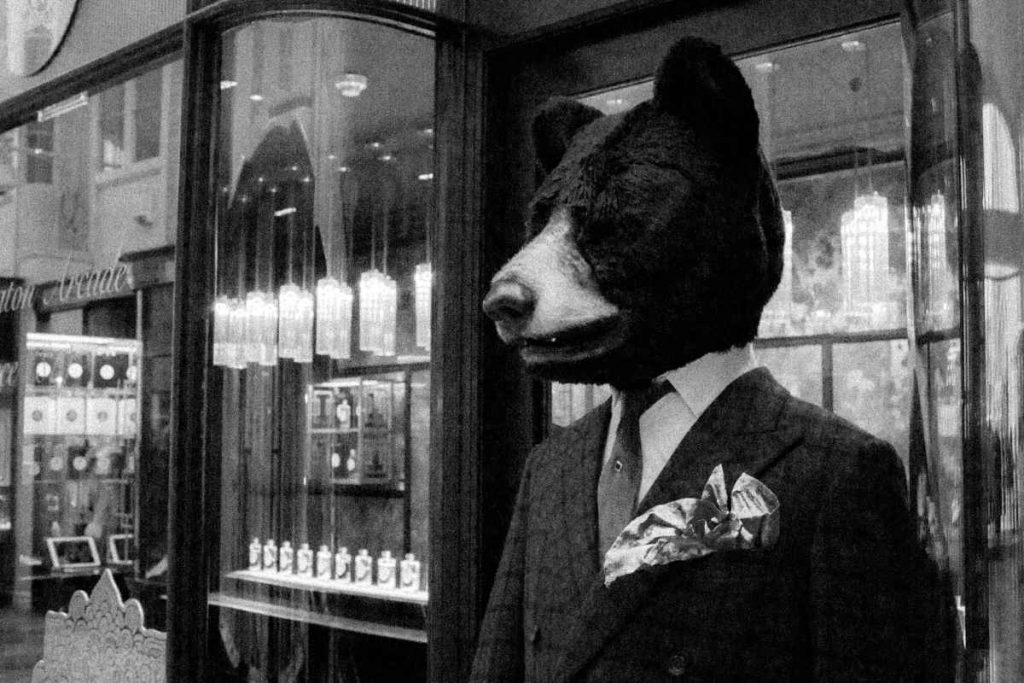 B&W picture of a man in a window pane suit while wearing a bear head mask