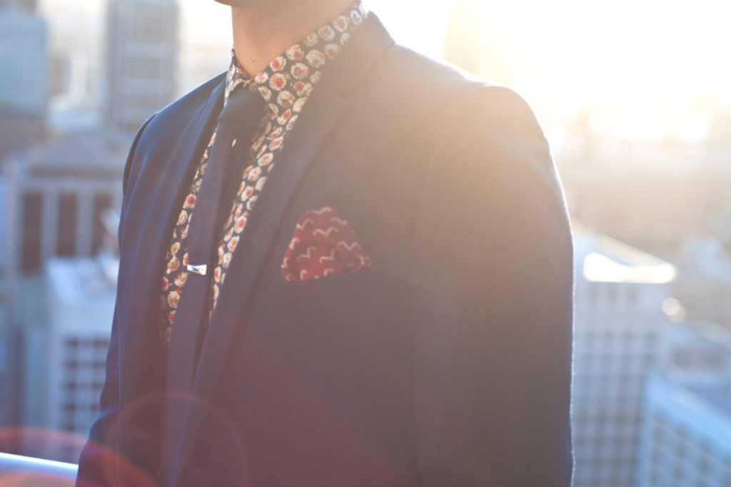 Close up of male upper torso wearing a geometric patterned shirt, blue tie and blue suit jacket with a red patterned pocket square