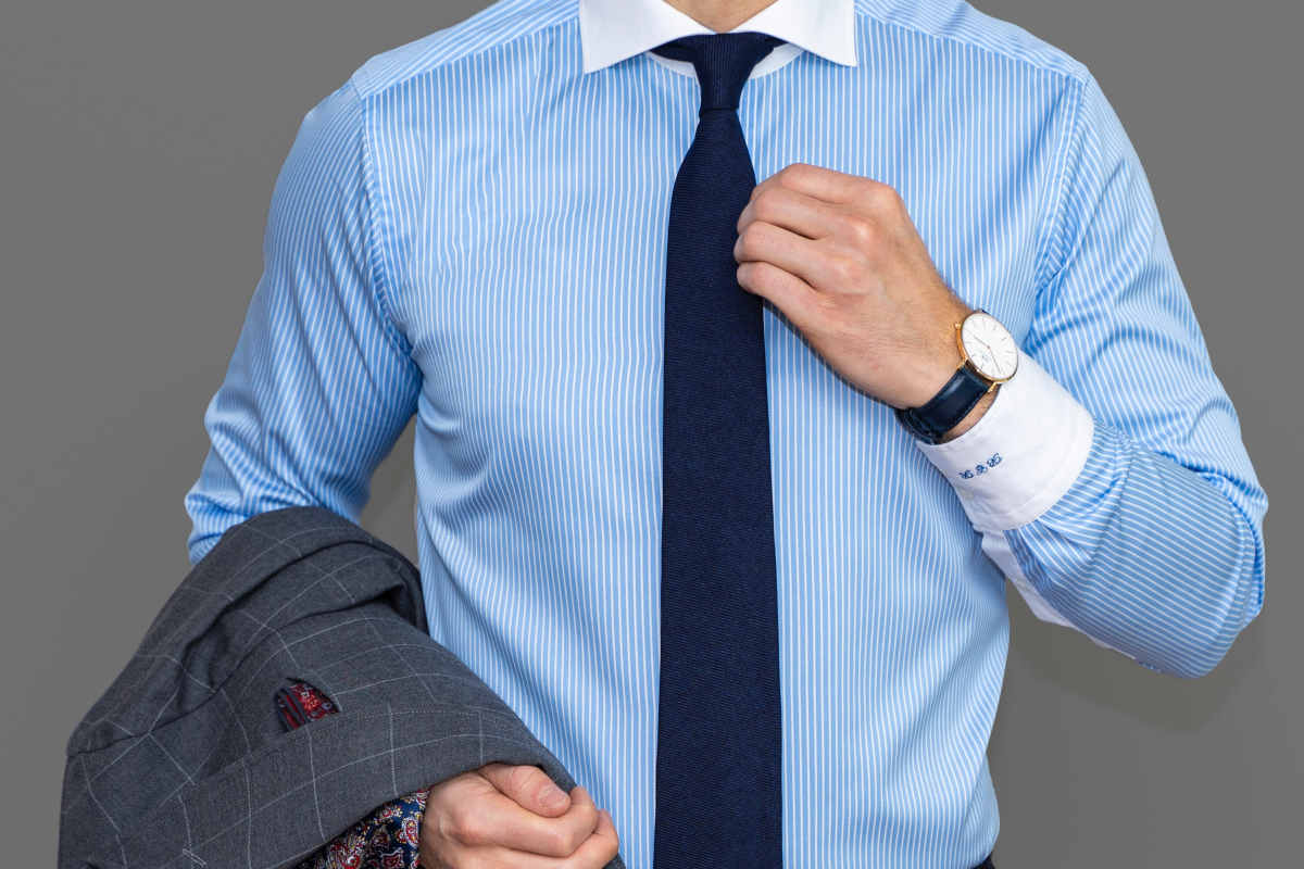 Man's torso wearing a blue striped Winchester shirt, navy jacquard tie and carrying a suit jacket on his arm