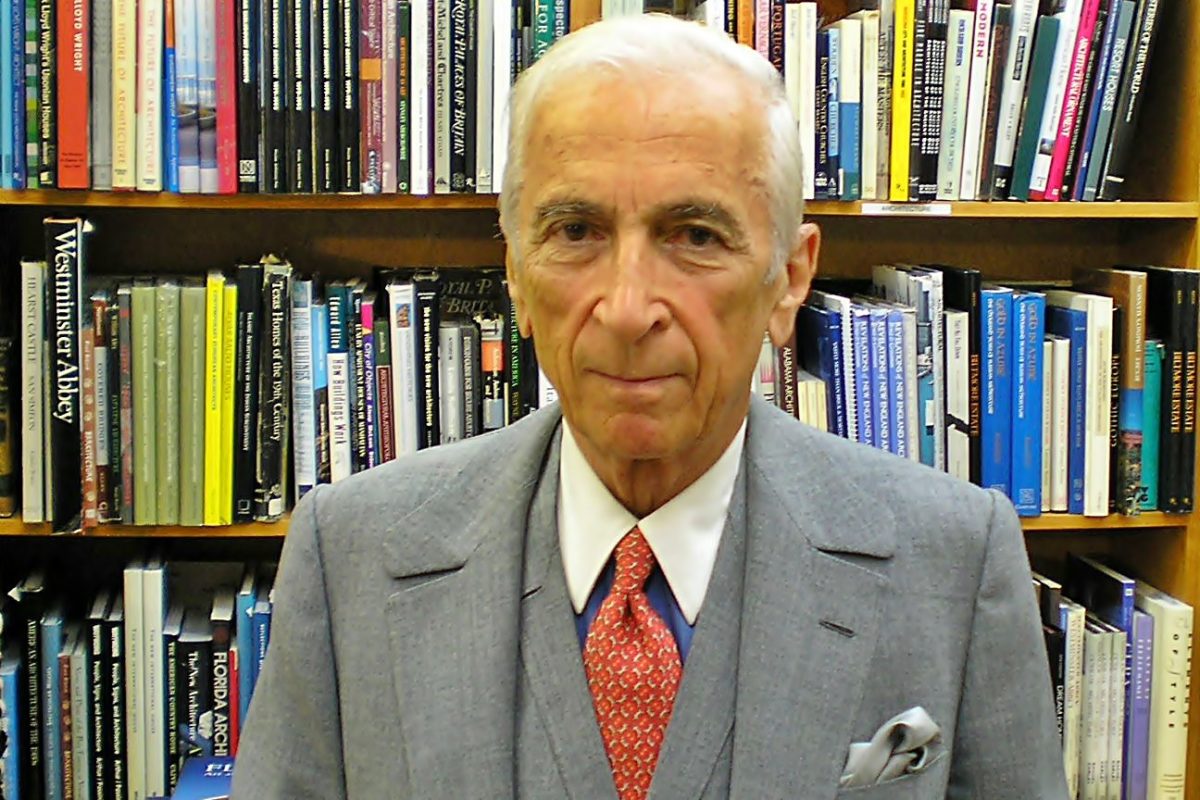 Gay Talese wearing a medium gray 3-piece suit over a royal blue winchester shirt, with a red-orange macclesfield tie and a silver pocket square, in front of a bookshelf