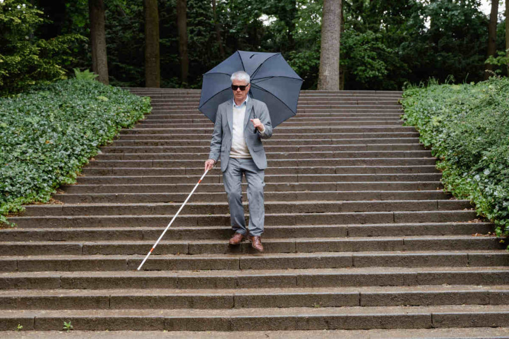 Blind man wearing a medium grey suit, brown derbies, a light blue shirt and a cream crew-neck sweater, walking down a park steps with a cane on one hand a an umbrella on the other