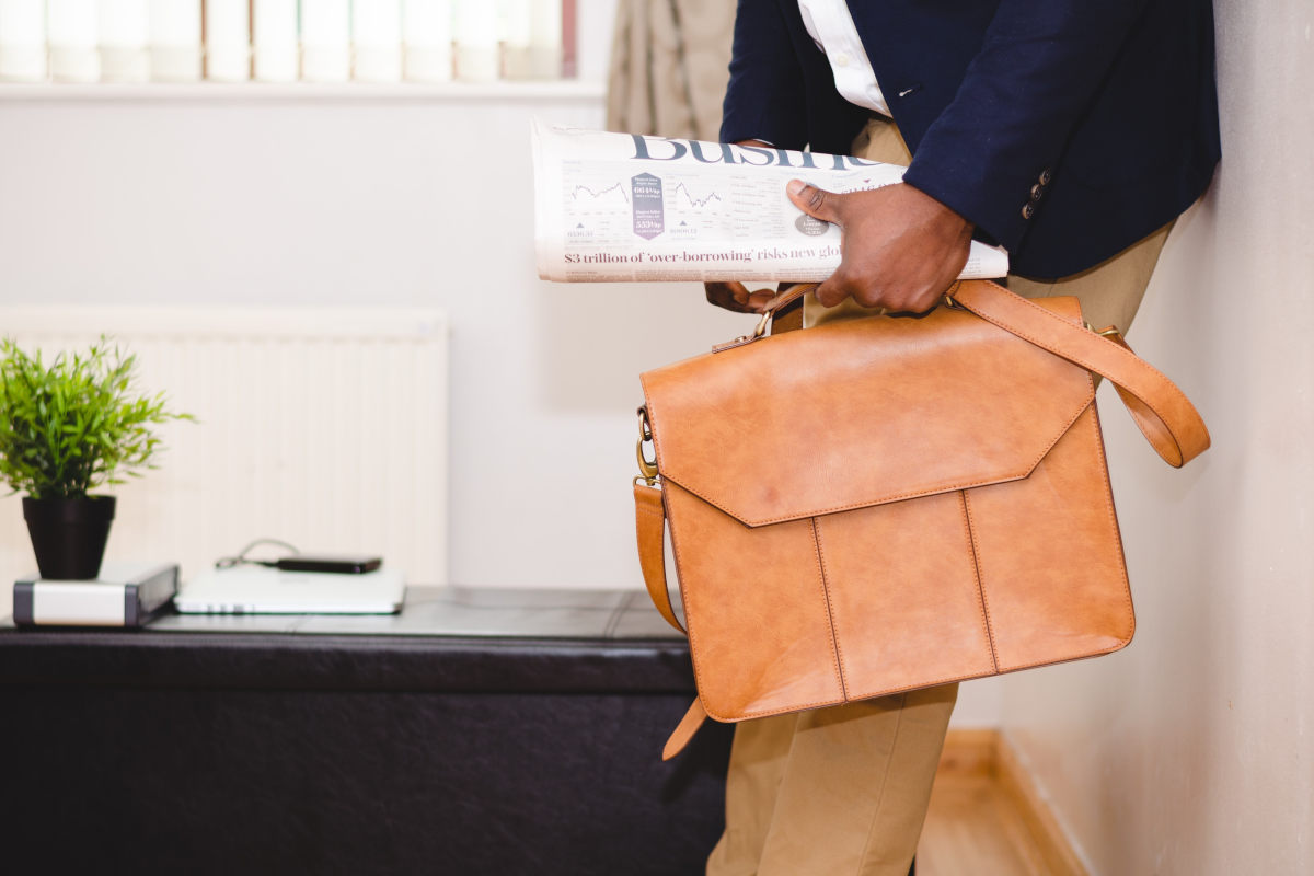 Waist-down shot of a man standing against an office wall, wearing tan chinos, white dress shirt and navy jacket, holding a brown briefcase and a folded newspaper