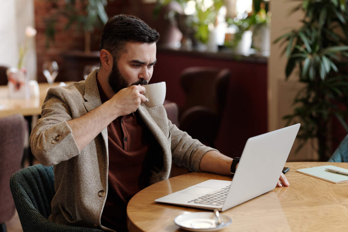 Man wearing a sand coloured wool jacket over a burgundy polo shirt, sipping coffee while working on a laptop computer
